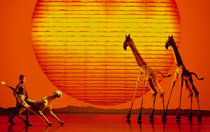 Schauspieler in Tierskostümen vor einer großen Sonne auf der Bühne.