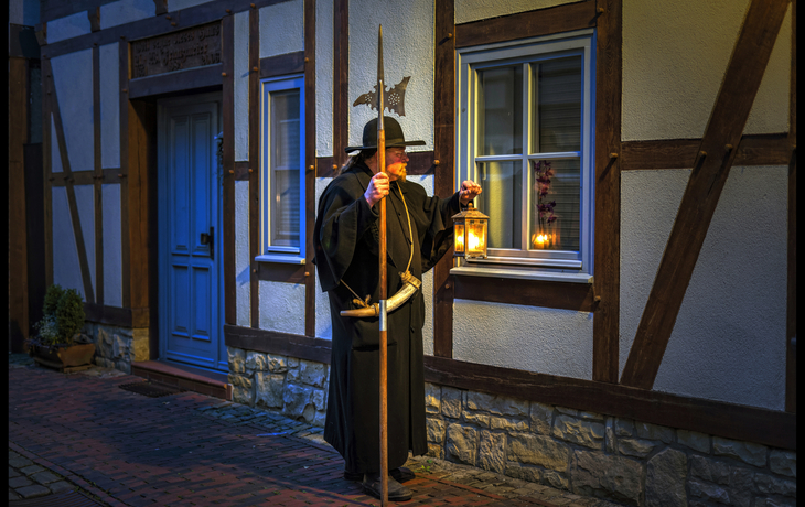 Ein mittelalterlicher Nachtwächter mit Hellebarde und Laterne vor Fachwerkhaus.