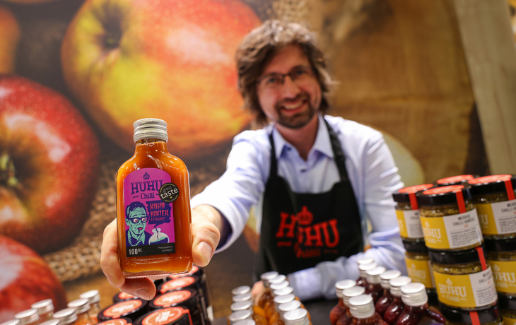 Mann präsentiert Huhu Chili-Soßenflasche auf einem Marktstand.