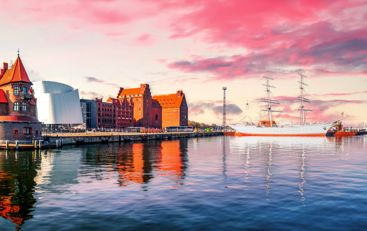 Hafenansicht mit historischen Gebäuden und Segelschiff bei Sonnenuntergang.
