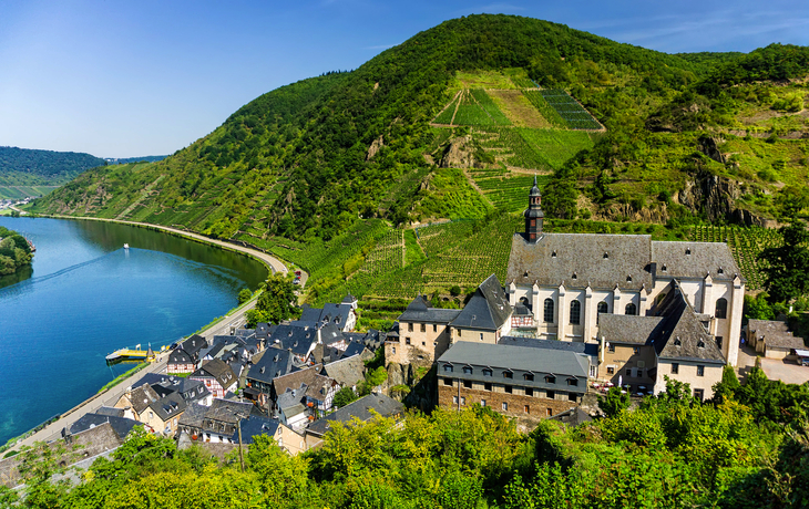 Mittelalterliches Dorf mit Kirche an einem Fluss in einer grünen Hügellandschaft.