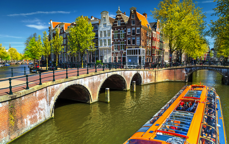 Gracht in Amsterdam mit Häusern und Touristenboot unter blauem Himmel.