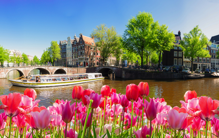 Kanal in Amsterdam mit Brücke, Tulpen und Boot bei blauem Himmel.