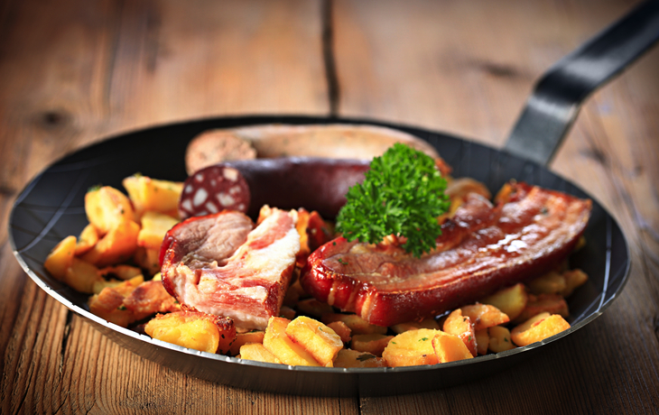 Pfanne mit verschiedenen Wurstsorten und Bratkartoffeln auf Holztisch.