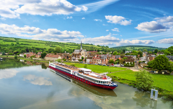 Flusskreuzfahrtschiff auf der Seine nahe eines Dorfes mit Hügeln im Hintergrund.