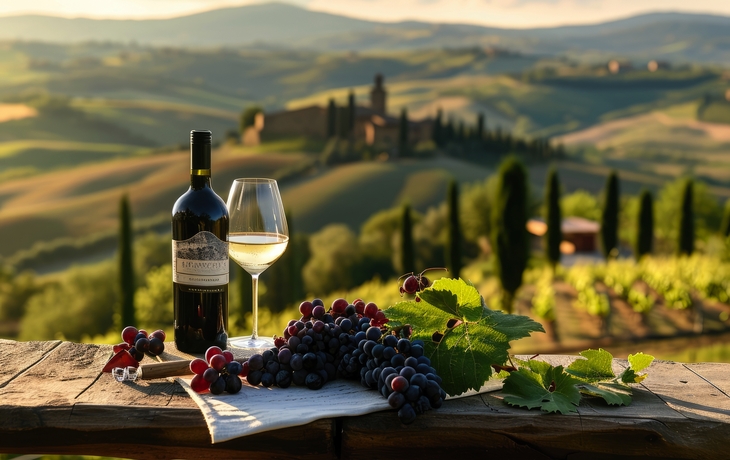 Weinflasche und Glas auf Tisch mit Weintrauben, im Hintergrund hügelige Landschaft.