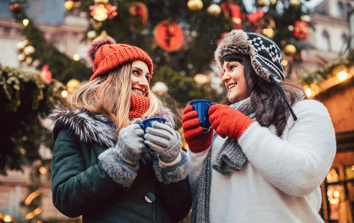 Zwei Frauen trinken Glühwein auf einem Weihnachtsmarkt vor einem geschmückten Baum.