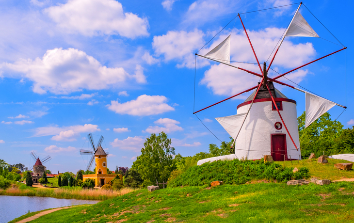 Drei Windmühlen auf einem grünen Hügel unter blauem Himmel mit Wolken.