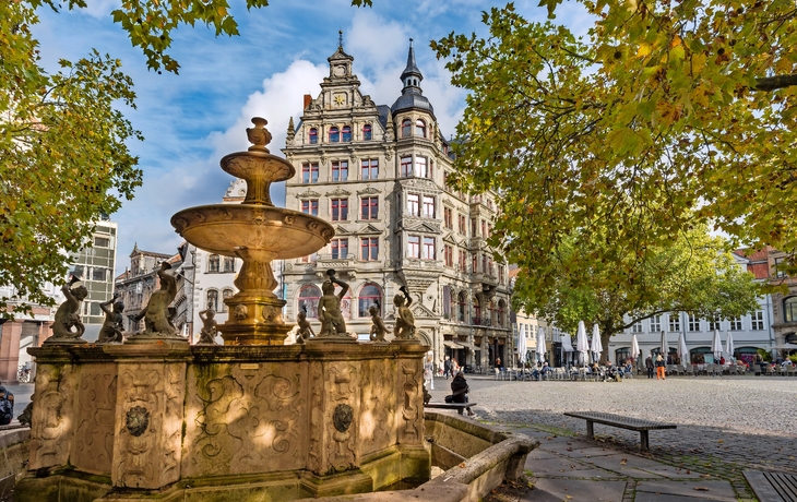 Historischer Brunnen und Gebäude an einem sonnigen Platz, umgeben von Herbstbäumen.