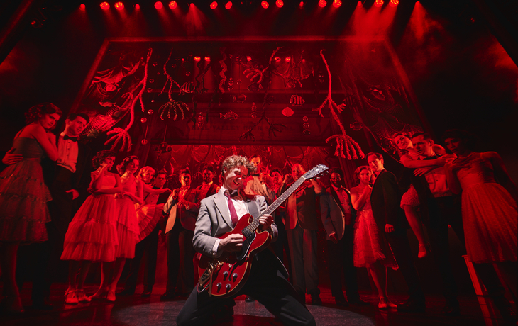 Mann spielt E-Gitarre auf Bühne mit rotem Licht, umgeben von Tänzern in eleganter Kleidung.