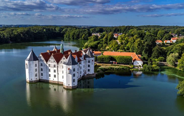 Weißes Schloss auf einer Insel in einem See, umgeben von Wald und blauen Himmel.