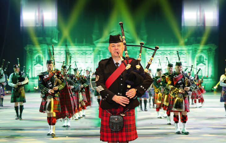 Ein Dudelsackspieler führt eine Parade in Kilts vor einer beleuchteten Kulisse an.