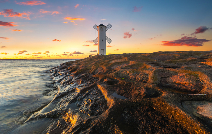 Leuchtturm bei Sonnenuntergang an felsiger Küste mit wolkigem Himmel.