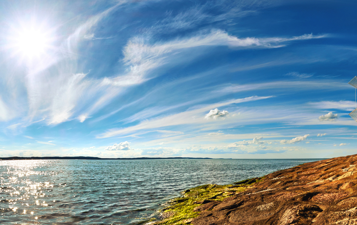Leuchtturm am Meeresufer unter blauem Himmel mit Wolken und Sonne