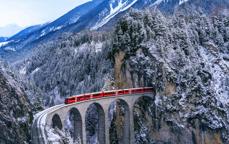 Roter Zug auf Viadukt in verschneiter Berglandschaft