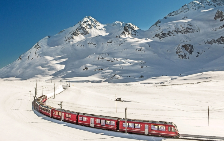 Roter Zug fährt durch verschneite Berglandschaft mit blauen Himmel.