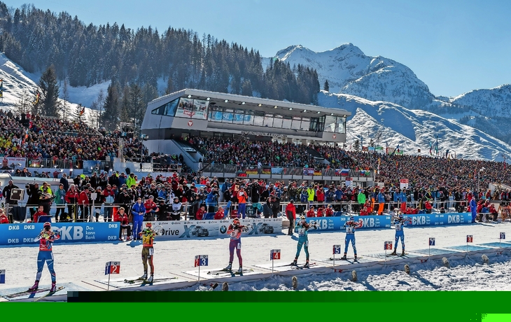 Blick auf ein vollbesetztes Biathlon-Stadion in verschneiter Berglandschaft.