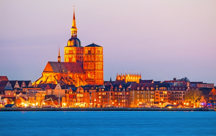 Stadtansicht mit beleuchteter Kirche bei Sonnenuntergang am Wasser.