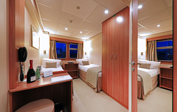 Kabine mit zwei Betten, Fenster, Schreibtisch und Schrank auf einem Schiff.