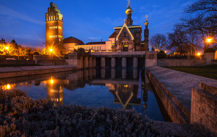 Historische russische Kapelle bei Nacht mit beleuchtetem Turm im Hintergrund.