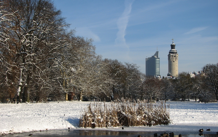 Verschneiter Park mit Teich, Enten und Stadtsilhouette im Hintergrund