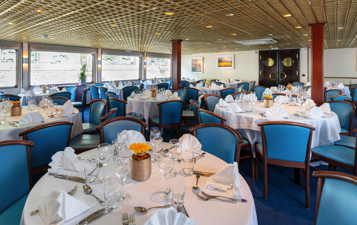 Elegantes Schiffsrestaurant mit gedeckten Tischen und blauen Stühlen.