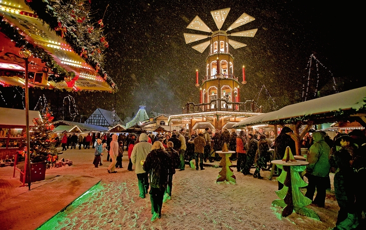 Menschenmenge auf beleuchtetem Weihnachtsmarkt bei Nacht, Schnee fällt.