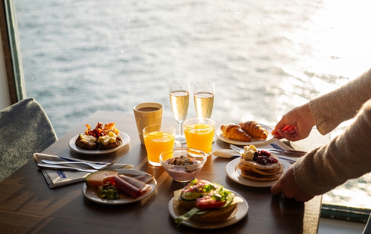 Frühstückstisch mit Gebäck, Saft, Kaffee und Sekt vor einem Schiffsfenster.