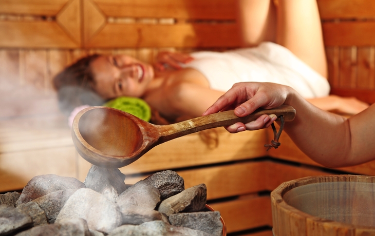 Frau entspannt in Sauna beim Aufguss, Hand hält Kelle über heißen Steinen.