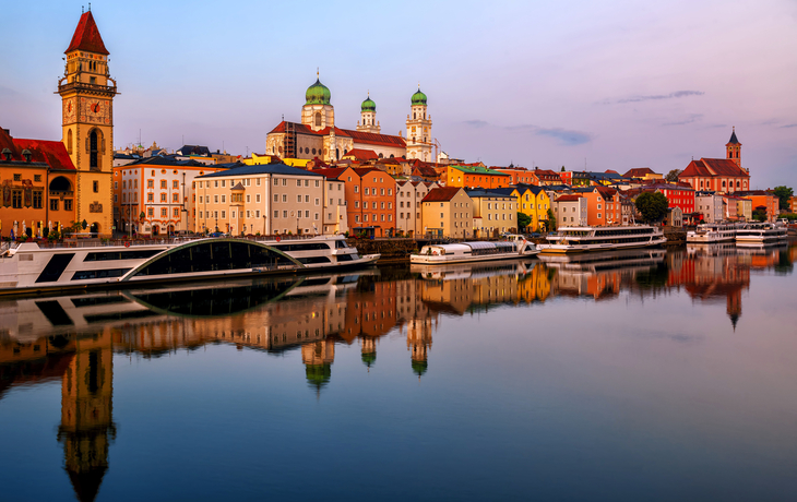 Flussufer mit Stadtansicht bei Sonnenuntergang, Schiffe und historische Gebäude im Hintergrund.