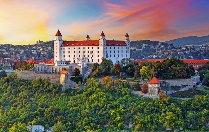 Burg auf einem Hügel mit rotem Dach vor buntem Sonnenuntergangshimmel.