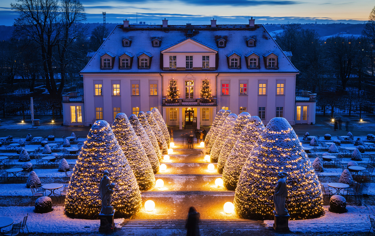 Beleuchtetes Herrenhaus im Winter mit schneebedecktem Garten bei Dämmerung.