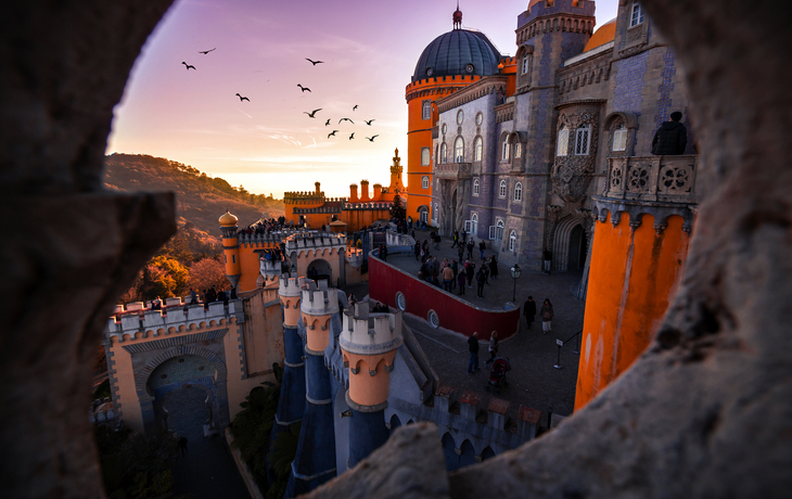 Blick auf buntes Schloss bei Sonnenuntergang, umgeben von Hügeln und Vögeln am Himmel.