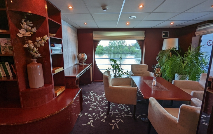 Bibliothek im Schiff mit Sesseln, Pflanzen, Bücherregal und großem runden Fenster.