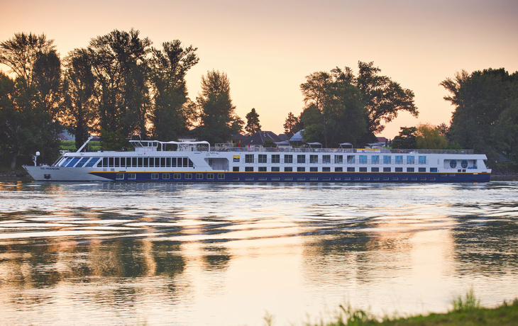 Flusskreuzfahrtschiff fährt bei Sonnenuntergang auf einem Fluss, umgeben von Bäumen.