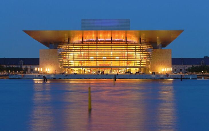 Ein beleuchtetes Gebäude mit modernem Design, das sich nachts am Wasser spiegelt.