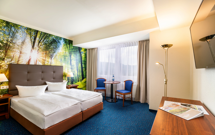 Hotelzimmer mit Doppelbett, Tisch, Stühlen und Waldtapete an der Wand.