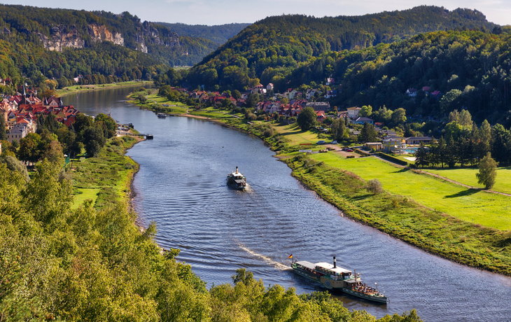 Flusslandschaft mit Hügeln, zwei Schiffen und Dörfern im Hintergrund.