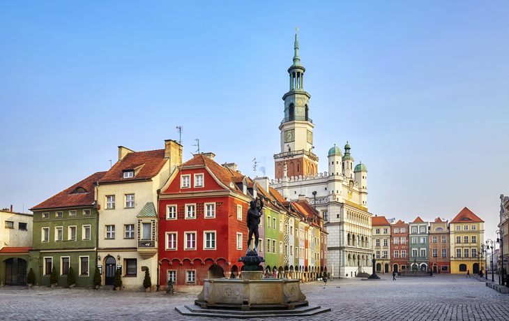 Bunte Häuser und Rathaus auf einem Platz mit Brunnen und Kopfsteinpflaster.