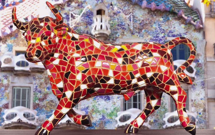 Stierfigur mit buntem Mosaikmuster vor verziertem Gebäude.