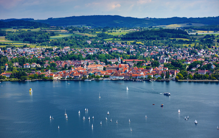 Kleinstadt am See mit Segelbooten und Hügeln im Hintergrund.