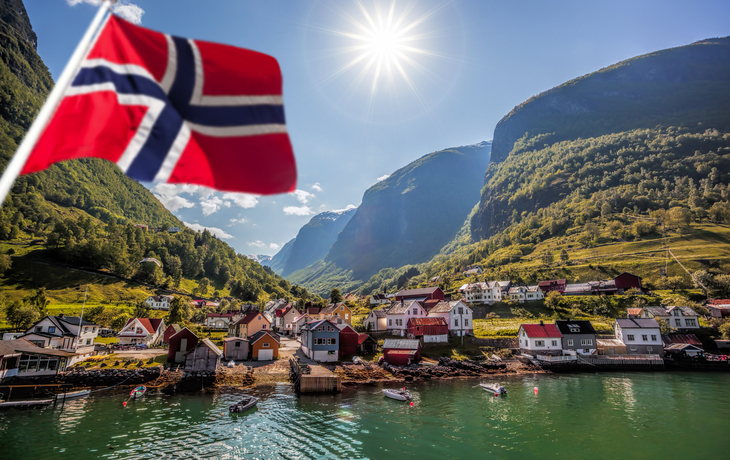 Norwegische Flagge vor Fjordlandschaft mit Dorf und Bergen bei Sonnenschein.