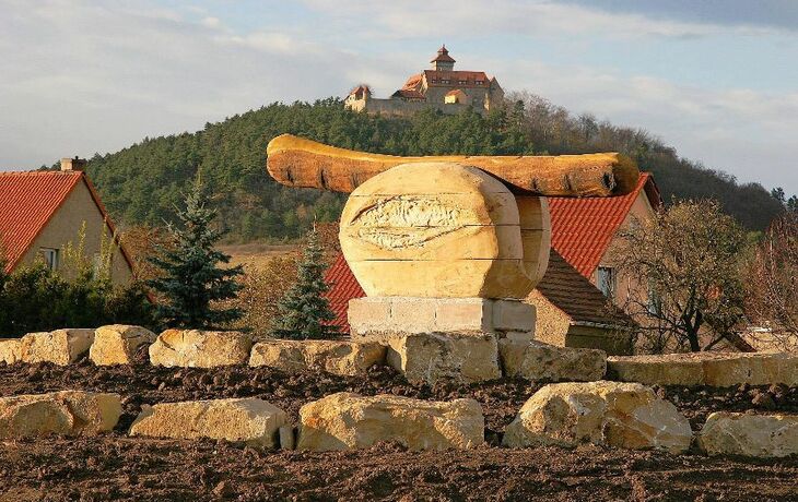 Holzskulptur einer Bratwurst auf einem Kreisverkehr, Hintergrund mit Hügel und Gebäude.