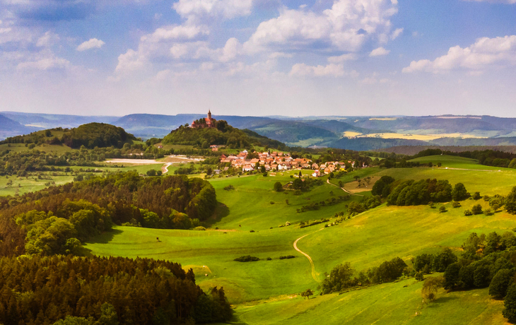 Landschaft mit Hügeln, kleinem Dorf und Burg im Hintergrund unter blauem Himmel.