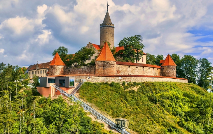 Historische Burg auf einem bewaldeten Hügel mit Zahnradbahn.